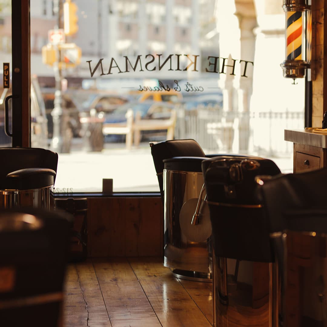 View from inside The Kinsman looking out onto a West Village street, featuring the logo on the window and classic leather barber chairs.