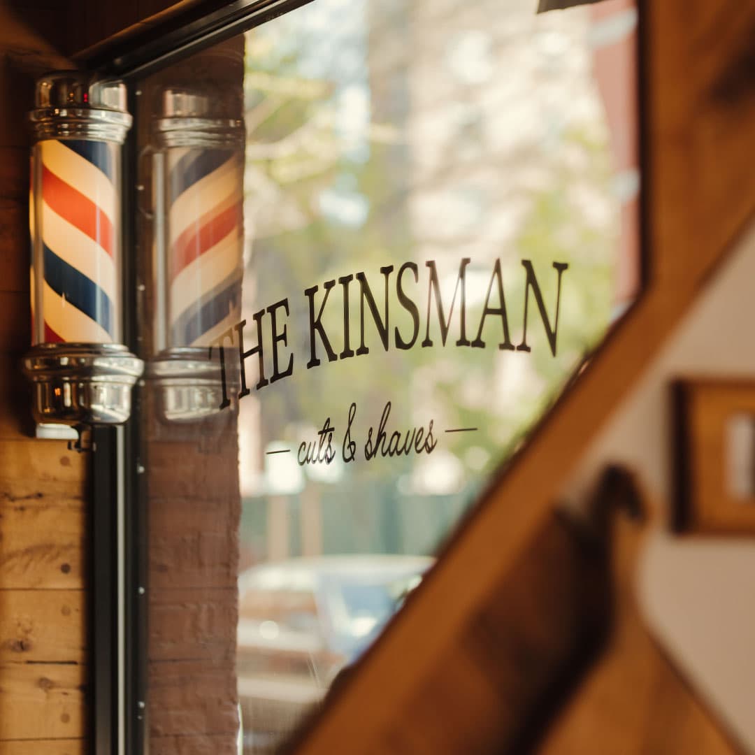 A traditional striped barber pole hangs outside The Kinsman barbershop on a brick building in New York City. The window sign reads "THE KINSMAN - cuts & shaves."