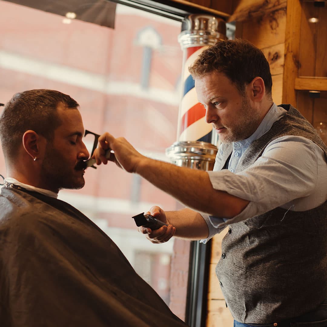 A barber using electric clippers and a comb to perform a precision skin fade haircut on a client in the West Village, NYC.