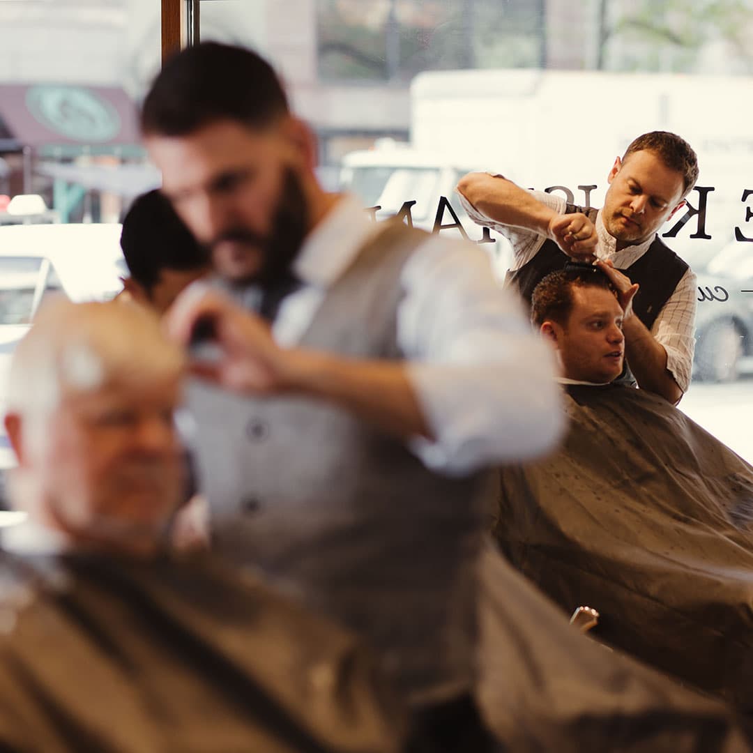 A vintage-style portrait view showing a master barber giving a classic taper haircut to a client at The Kinsman shop in NYC.