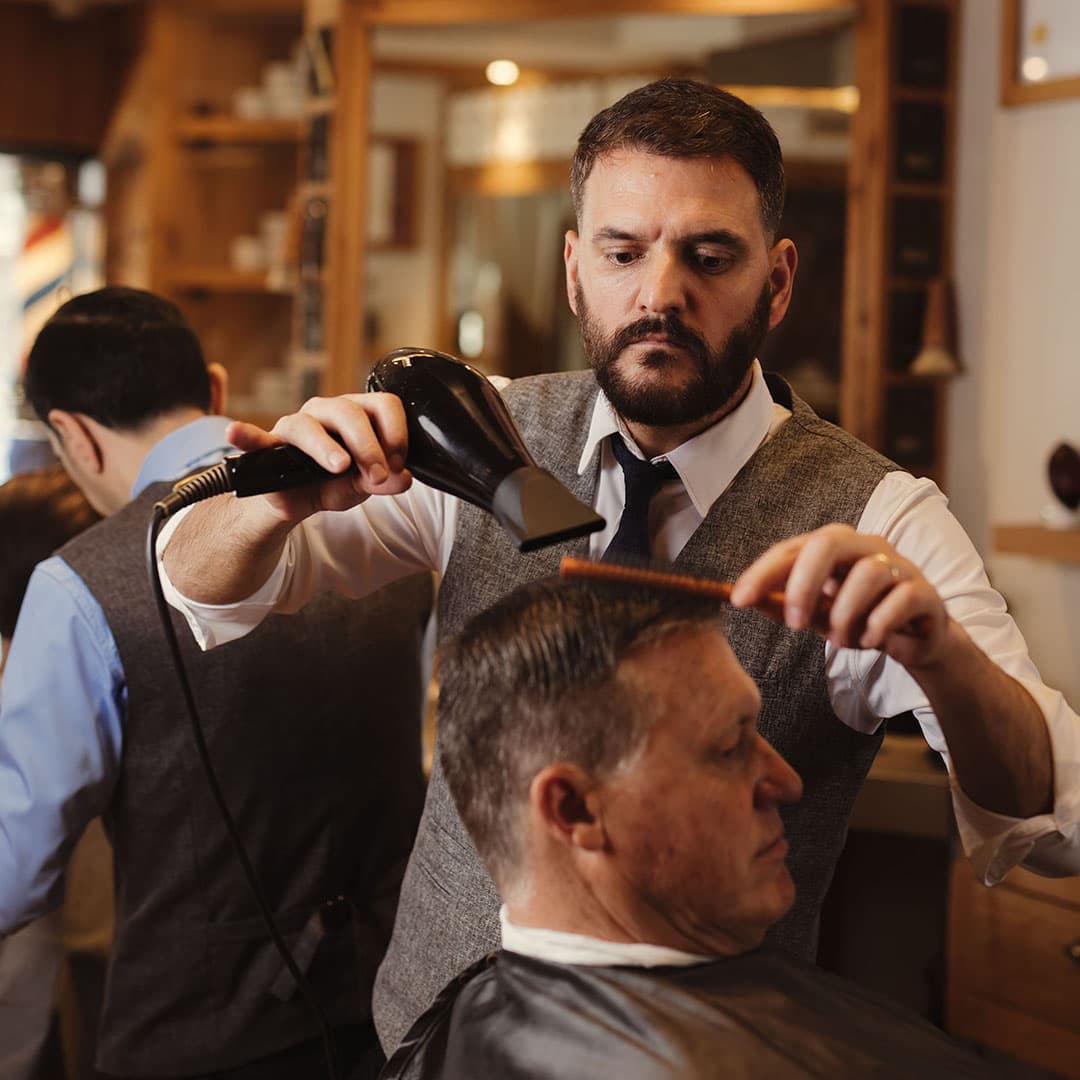 A barber using a blow dryer and brush to professionally style a client's hair after a cut, illustrating the finished, professional style.