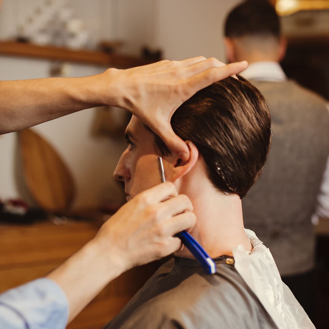 Detail shot of a barber using a straight razor to clean up the neckline and sideburns for a sharp, modern men's haircut in NYC.