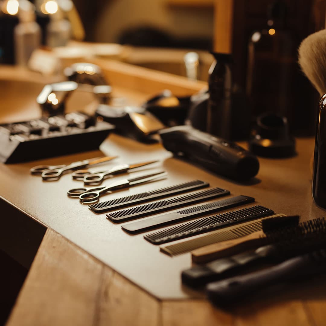 An organized row of professional barber shears, combs, and hair clippers on a wooden workstation, showcasing the high-end equipment at The Kinsman.