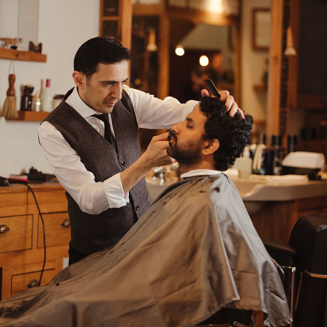 A detailed, focused portrait of a barber performing a detailed mustache trim on a client with dark, curly hair.