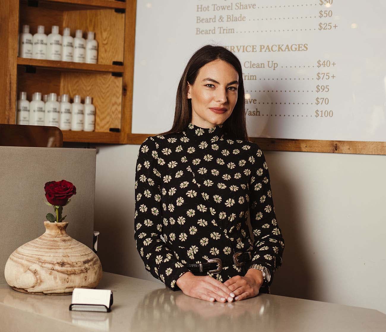 Portrait of The Kinsman reception desk staff in the West Village, with a visible price list featuring services like hot towel shave, regular cut, and skin fade, adjacent to product shelves.