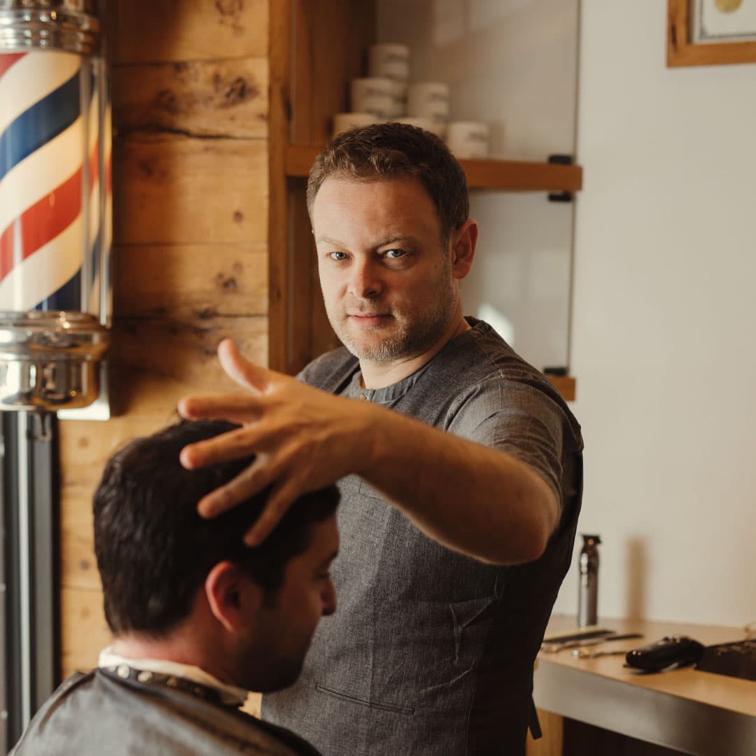 Portrait of an expert NYC barber finishing a haircut, with a traditional barber pole and wood-grain interior providing a classic atmosphere.