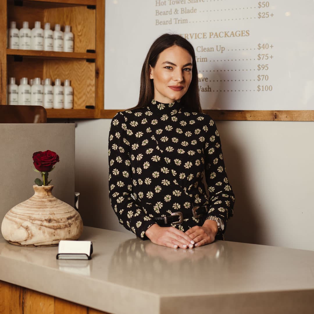 A welcoming female receptionist standing at the front desk of The Kinsman barbershop in the West Village, with a prices list and red rose vase visible.