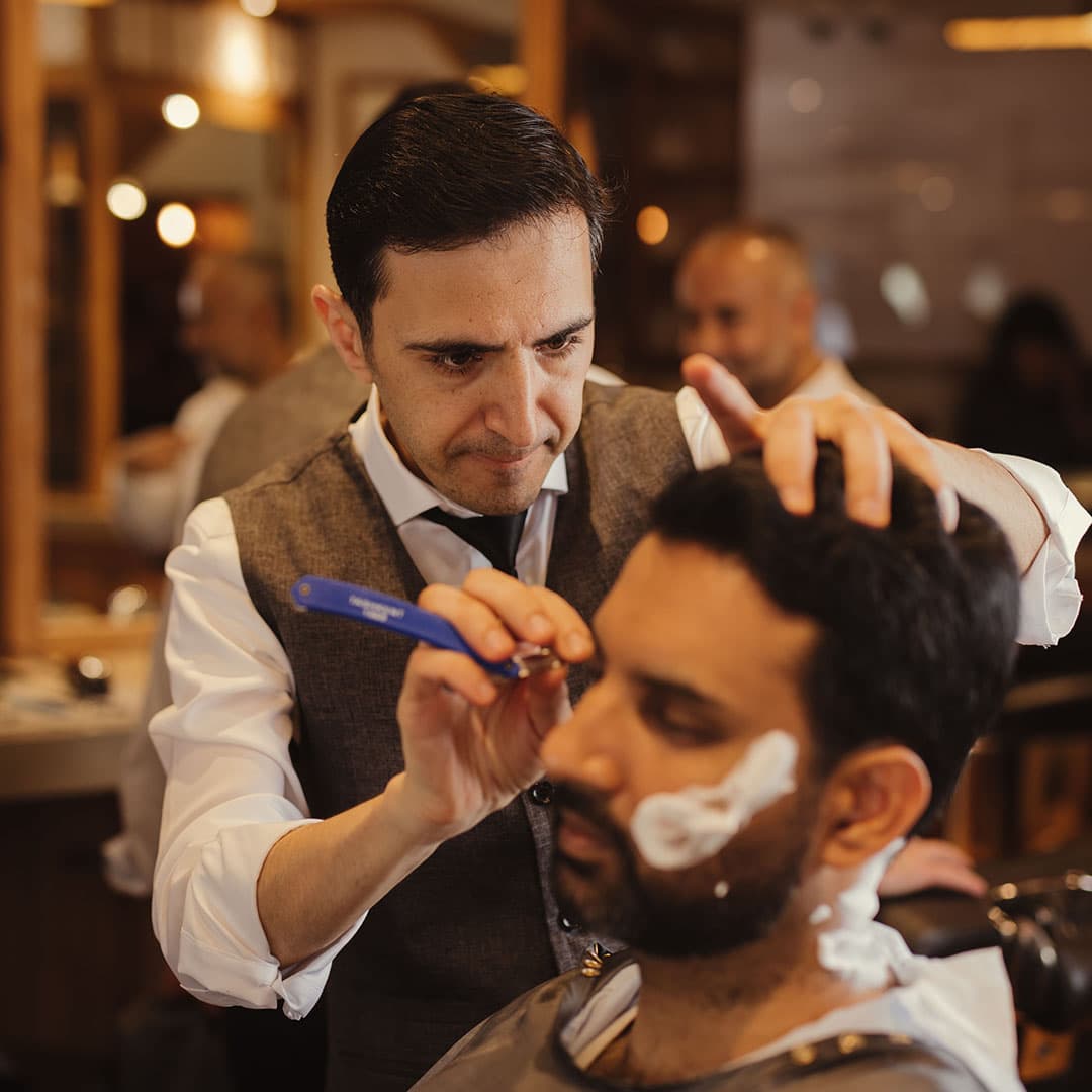 Close-up action shot of a barber using a straight razor for a traditional hot towel shave experience, showing the shave cream and razor angle.